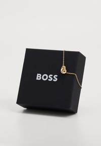 Zlatna ogrlica s teksturiranim ovalnim privjeskom, prikazana naspram crne kutije s kontrastnim bijelim logotipom "BOSS".
