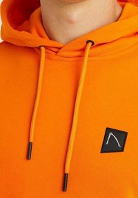 Felpa arancione brillante con coulisse e una piccola toppa quadrata nera con un design astratto di linee bianche sul petto.