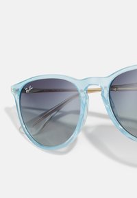 Ochelari de soare transparenți de culoare albastru deschis, cu lentile gri degradé și accente metalice aurii pe punte și pe mâini. Design curat, modern.