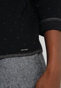 Maglione di maglia nera con riflessi metallici, scollatura a barchetta e maniche corte. Rifinito con un orlo glitterato e un piccolo etichetta con il marchio.