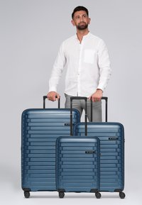 Tre valigie resistenti di colore blu di diverse dimensioni, con strisce testurizzate e maniglie nere estraibili, poggiate su quattro ruote.