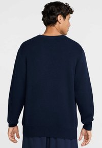 Ναυτικό μπλε πλεκτό πουλόβερ με στρογγυλή λαιμόκοψη, μακριά μανίκια και ριμπ μανσέτες. Η πίσω όψη δείχνει χαλαρή εφαρμογή και υφασμάτινη υφή.