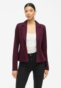 Burgundský elegantní blazer s klopovým límcem, zapínáním na jeden knoflík a dlouhými rukávy, kombinovaný s bílým topem a černými džíny.