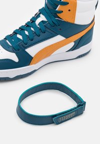 Bílé a tyrkysové kotníkové tenisky s oranžovým pruhem, tyrkysovými tkaničkami a odpovídajícím tyrkysovým páskem kolem kotníku s logem "PUMA" na bílém pozadí.