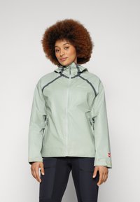 Veste imperméable vert clair avec capuche, présentant des accents gris, des poches zippées et des poignets ajustables. Texture lisse, design fonctionnel.