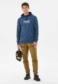 Niebieska bluza z kapturem z kieszenią z przodu i logo, w połączeniu z brązowymi spodniami do wspinaczki z czarnymi akcentami. Model trzyma buty wspinaczkowe i ma na sobie czarne sneakersy.