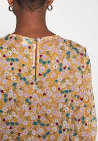 Kurze, lockige Haare und goldene Creolen, trägt eine senfgelbe Bluse mit floralem Muster in Pink, Weiß und Türkis, mit Knöpfen am Rücken nahe dem Hals.