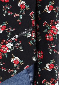 Tissu à motifs floraux noirs, avec des fleurs rouges et blanches. Comprend un détail de fermeture éclair argentée sur le côté. Matière douce et texturée.