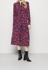 Frau trägt ein langärmeliges, knielanges Kleid mit pink-rot floralem Muster, kombiniert mit schwarzen, hochhackigen Stiefeletten, vor einem weißen Hintergrund stehend.