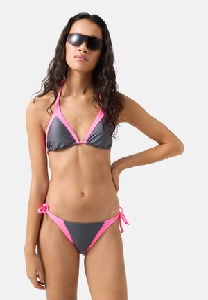Woman wearing gray and pink bikini and large black sunglasses, standing with one hand on her hip against a plain white background.