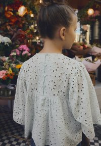 Haut en coton blanc avec broderie en œillet, manches bouffantes et taille froncée, avec un simple détail de bouton à l'arrière.