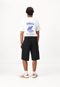 Hvit T-skjorte med blå grafisk trykk på baksiden, paired med svarte denimshorts og hvite sneakers med røde detaljer.