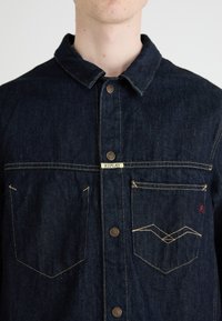Mørk denimjakke med krage, frontlukkning med knapper, to brystlommer med kontrasterende søm og en liten logoetikett.