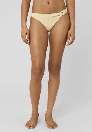 Femme portant un bas de bikini beige avec un détail d'anneau latéral, debout pieds nus sur un fond gris clair uni.