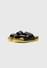 Melni sandales ar divām regulējamām siksnām, korķa zoli un spilgti zaļu gumijas zoli, ar atvērtu purngala dizainu.