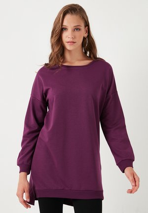 Purple long-sleeve sweatshirt made of soft fabric, featuring a wide neckline, dropped shoulders, and a longer rear hem. Subtle side slits.