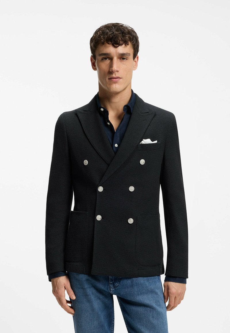 Jeune homme portant un blazer croisé sombre avec des boutons blancs, une chemise bleu marine, un mouchoir blanc dans la poche et un jean bleu, debout devant un fond blanc.