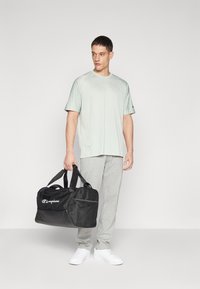 Camiseta deportiva verde claro, pantalones de chándal grises, zapatillas blancas y una bolsa de deporte negra con un bolsillo de malla, que presenta el logo blanco de "Champion".