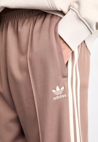 Bruna sportbyxor med vita sidosträck och en broderad logotyp. Har ett mjukt, texturerat tyg och en elastisk midja med fickor.