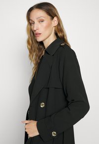 Svart trenchcoat med dobbeltradet design, gullfargede knapper og bred krage. Glatt stoff med en flytende silhuett.