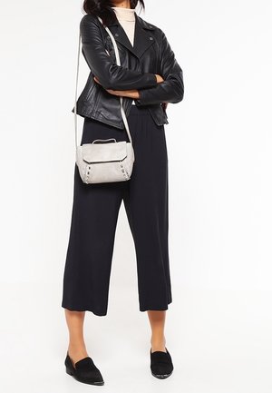 Femme portant une veste en cuir noire, un pantalon ample noir, un col roulé beige, des mocassins noirs, portant un sac bandoulière gris clair, les bras croisés.