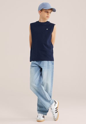 Garçon portant un débardeur bleu marine, un jean bleu clair, des baskets blanches et une casquette bleu clair, debout, les bras derrière le dos.