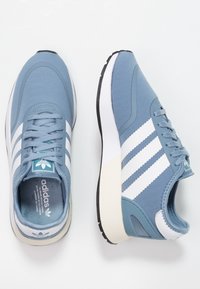 Ljusblå sneakers med vita ränder, textilöverdel, rundad tå, vadderad sula och en logotypetikett på tungan. Texturerade och släta ytor.