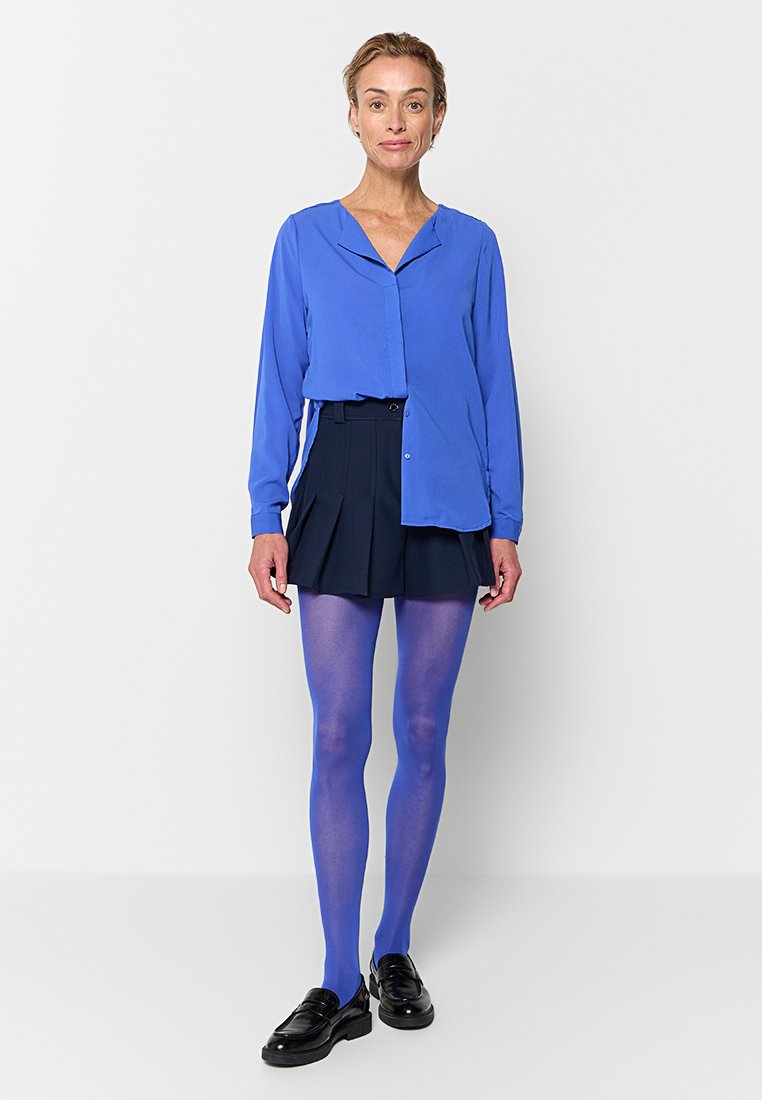Langärmlige blaue Bluse mit Kragen, kombiniert mit einem plissierten marineblauen Rock und blickdichten blauen Strumpfhosen. Schwarze Slipper runden das Outfit ab.