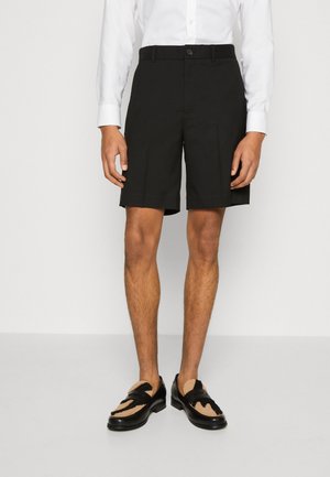 Schwarze maßgeschneiderte Shorts mit flacher Vorderseite, ausgestattet mit einem Knopfverschluss und Seitentaschen, kombiniert mit geflochtenen Sandalen. Beine und Füße sind sichtbar.