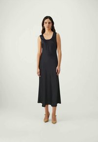 Vestido maxi preto, sem mangas, com um tecido liso e elástico, apresentando um decote redondo e uma bainha fluída. Usado com sapatos de salto alto bege.