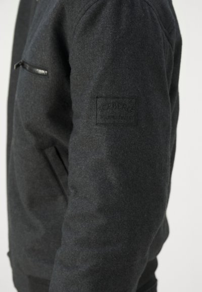 Chaqueta bomber negra con un acabado texturizado, que presenta un bolsillo lateral con cremallera y un parche con logo en relieve en la manga izquierda.