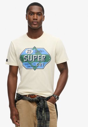 Kremowa koszulka z krótkim rękawem z niebieską i zieloną grafiką z napisem "SUPER DRY". Logo umieszczone na środku. Noszona z koszulą w kratę zawiązaną w talii.