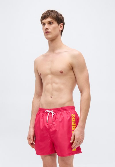 Νεαρός άνδρας όρθιος, φορώντας λαμπερά κόκκινα μαγιό με κίτρινα γράμματα «DSQUARED2» στο αριστερό πόδι και λευκό κορδόνι.