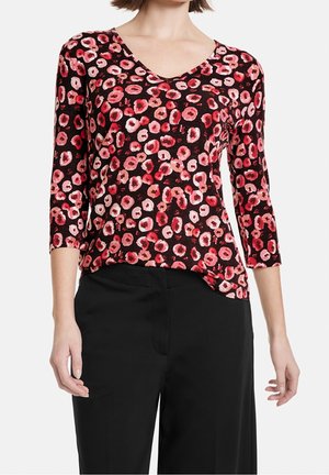 Femme portant un chemisier noir ajusté avec des motifs floraux circulaires roses et rouges et des manches trois-quarts, associé à un pantalon noir.