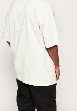 Persona che indossa una maglietta bianca a maniche corte oversize e pantaloncini neri, mostrata di spalle su uno sfondo semplice.