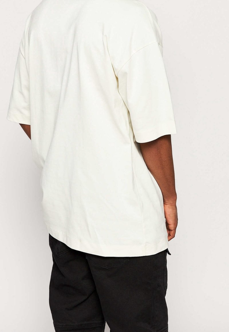 Personne portant une chemise blanche unie à manches courtes oversize et un short noir, vue de dos sur un fond uni.