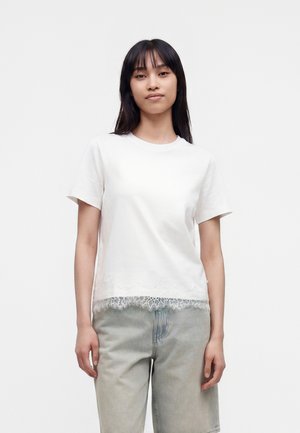 Jeune femme aux longs cheveux noirs portant un haut blanc uni à manches courtes avec une bordure en dentelle et un jean taille haute gris clair sur fond blanc.