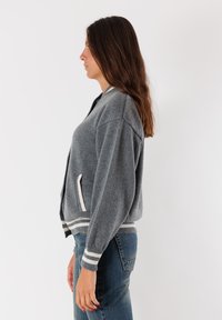 Donna con lunghi capelli castani, che indossa un cardigan grigio a maglia con strisce bianche e jeans blu, in posa di profilo contro uno sfondo bianco.