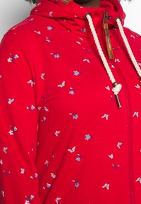 Czerwona bluza z kapturem na zamek, wykonana z miękkiego materiału, ozdobiona małym wzorem kwiatowym w kolorze niebieskim i białym. Posiada kaptur ze sznurkiem oraz ozdobne elementy na zamku.