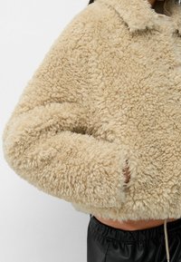 Beige kort faux-sheepskin-jakke med en tekstureret, fluffy overflade og en krave-udskæring. Har sidelommer og snøre detalje i kanten.
