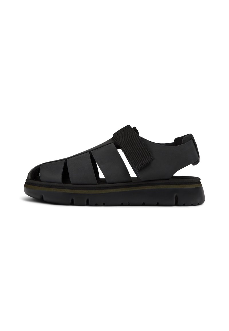 Black sandal with cutout design, adjustable velcro strap, thick sole with grooves, and a smooth texture. Features a subtle accent line.