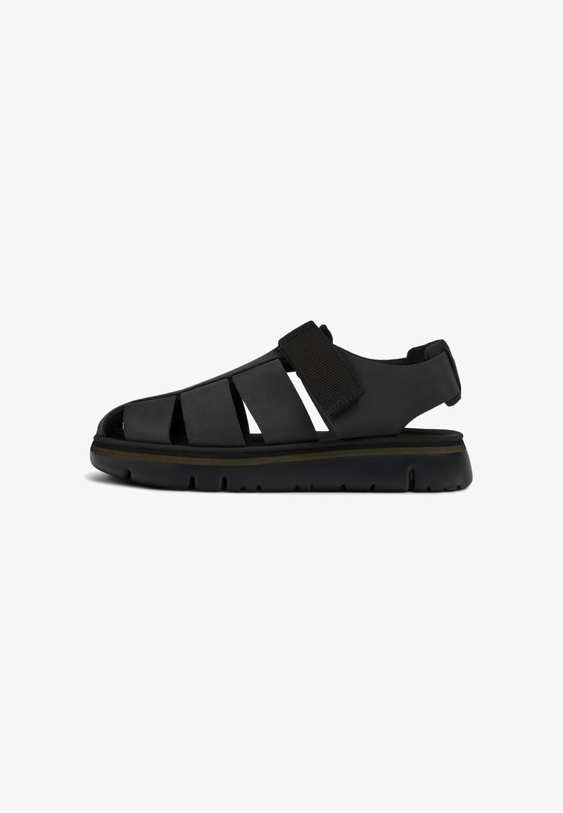 Black sandal with cutout design, adjustable velcro strap, thick sole with grooves, and a smooth texture. Features a subtle accent line.