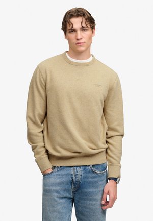 Junger Mann mit welligem Haar, der einen beigen Pullover und blaue Jeans trägt, mit einer Hand in der Tasche vor einem einfarbigen weißen Hintergrund.