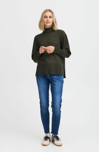 Oliwkowy sweter z golfem, z bocznymi rozcięciami, w połączeniu z niebieskimi rurkami i beżowymi trampkami z czarnymi sznurowadłami i akcentami.