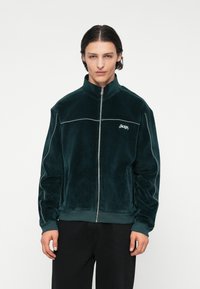 Tmavozelená zamatová bunda na zips s vysokým golierom, kontrastným bielym lemovaním a logom na ľavej hrudi. Model má na sebe čierne nohavice.
