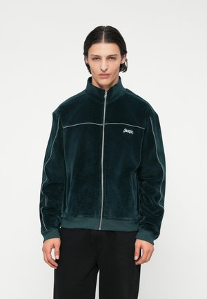 Giacca zip-up in velluto verde scuro con colletto alto, piping bianco a contrasto e logo sul petto sinistro. Il modello indossa pantaloni neri.
