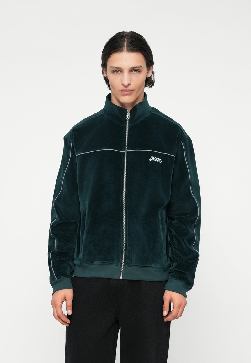 Tmavozelená zamatová bunda na zips s vysokým golierom, kontrastným bielym lemovaním a logom na ľavej hrudi. Model má na sebe čierne nohavice.