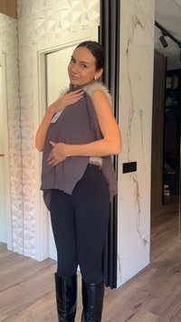 Γυναίκα με μαύρο παντελόνι και μπότες κρατάει ένα γκρι γιλέκο με γούνινη επένδυση σε ένα μοντέρνο δωμάτιο με υφασμένους λευκούς τοίχους και ξύλινο πάτωμα.