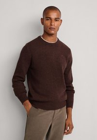 Mann trägt einen dunkelbraunen Rundhals-Pullover und beigefarbene Hose, steht mit einer Hand in der Tasche vor einem schlichten hellen Hintergrund.