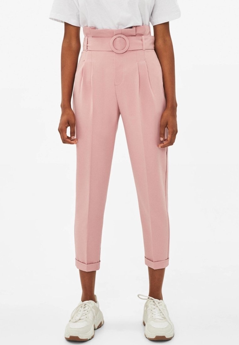 Personne portant un pantalon rose taille haute ceinturé avec des ourlets retroussés, associé à des baskets blanches et un haut blanc à manches courtes.